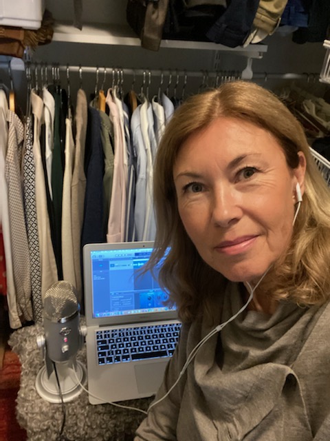 Bilden visar Åsa Melin som sitter i sin garderob efter att ha spelat in ett avsnitt av sin podd Klimakteriepodden. Åsa kollar in i kameran. Bakom henne syns en rad med upphängda skjortor, samt en laptop med en mikrofon bredvid sig.