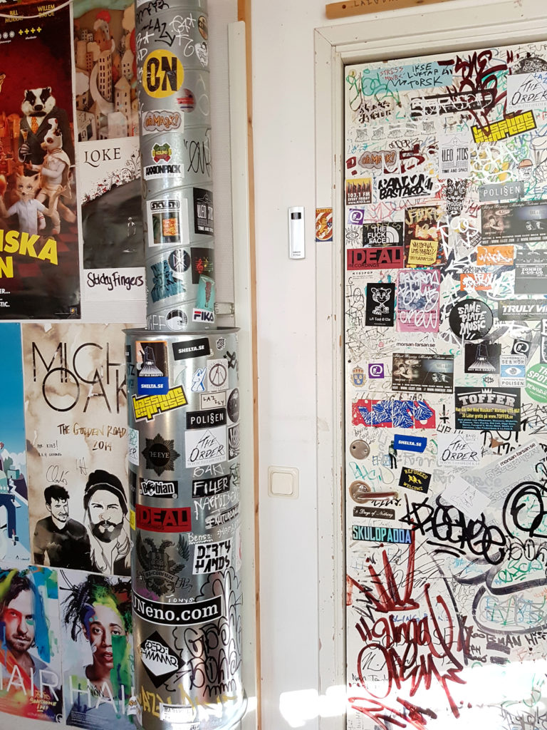 Bilden visar en bit av studions vägg. Överallt sitter klistermärken i olika färger och affischer från olika event och filmer.