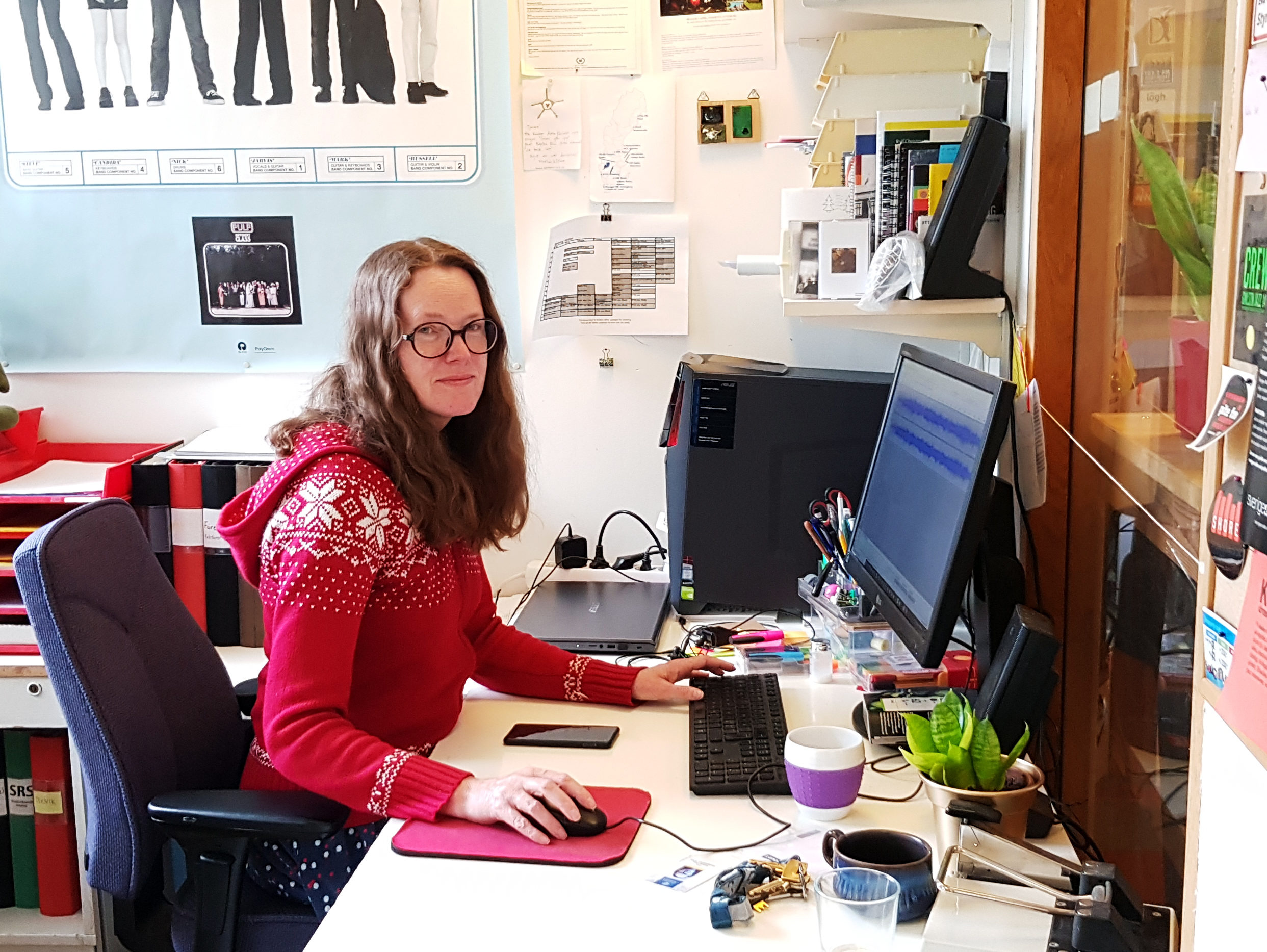 På bilden syns Eva Gustavfsson, som har en röd tröja och sitter vid ett skrivbord i sitt kontor. På skärmen kan man se att hon klipper ett radioinslag. 